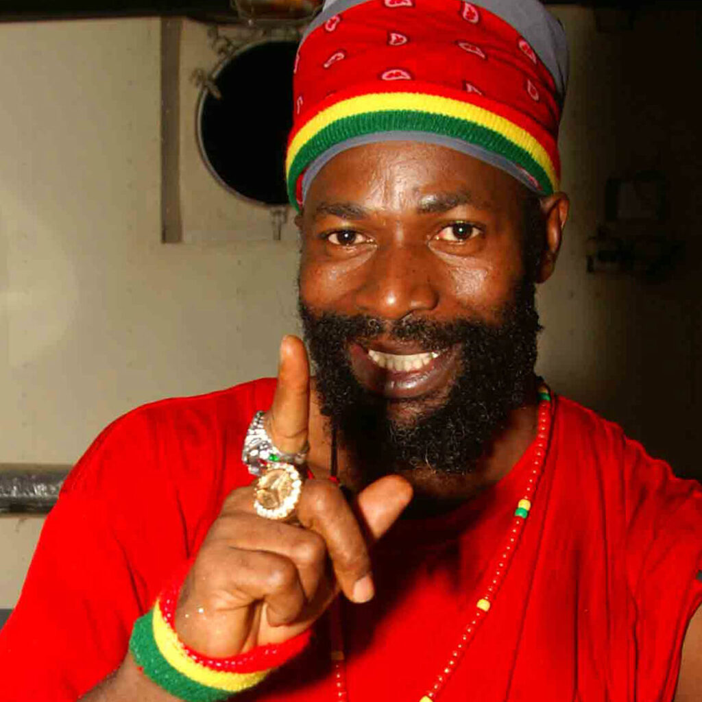 Listen to reggae and urban music at Cat Radio A smiling man with a beard, resembling Capleton, wears a red shirt and a red head wrap with green, yellow, and black stripes. He points upward and sports colorful beaded jewelry and rings. Cat Radio UK the number radio station